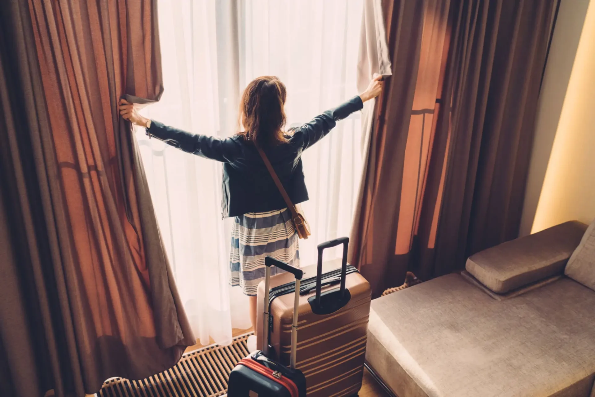 Person opening curtains in a hotel room with luggage.